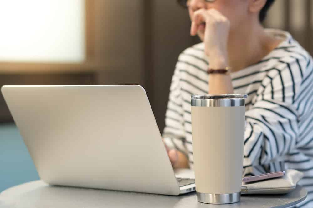 Smart looking woman use laptop computer to work from home due to Covid-19 pandemic, city lockdown and social distancing with her drink in a reusable stainless steel tumbler mug aside.